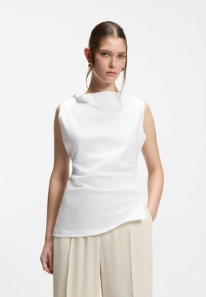 Kvinde med tilbagebundet hår iført hvid ærmeløs top og beige bukser med vide ben, stående med den ene hånd i lommen mod en hvid baggrund.