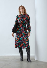 Blomstrete midikjole med svart bakgrunn, prydet med levende røde, lilla og grønne mønstre. Lange ermer og innsnevret midje, komplementert med svarte støvler.