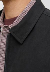Giacca nera intrecciata con chiusura a zip e colletto arrotondato, indossata sopra una camicia bordeaux con texture e motivo a quadri.