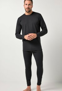 Hombre vestido con una camiseta ajustada de manga larga negra y mallas, de pie descalzo contra un fondo claro y liso, con las manos juntas cerca de la cintura.