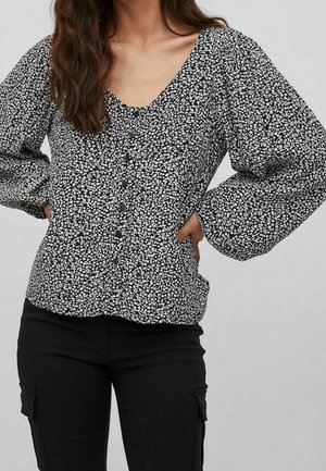 Frau trägt eine schwarz-weiß geblümte, langärmlige Bluse mit Knöpfen und schwarze Cargohosen, Hände in die Hüften gestützt.