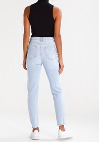 Persona vestida con jeans de tiro alto de color azul claro, top negro sin mangas y zapatillas blancas, de pie de espaldas sobre un fondo blanco.