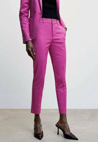 Pantalones rosas ajustados con una textura suave, que cuentan con un cierre de botón en la parte delantera y bolsillos, combinados con elegantes tacones negros de punta afilada.