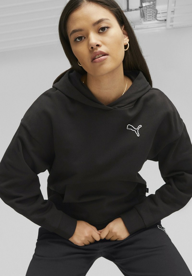 Felpa nera con cappuccio e logo Puma bianco sul petto, con una vestibilità comoda e tessuto testurizzato. Indossata con pantaloni neri abbinati.
