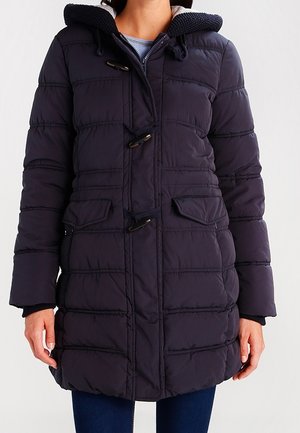 Vrouw die een donkerblauwe, halflange gewatteerde winterjas draagt met klepsluitingen en klepzakken, staand tegen een effen achtergrond.