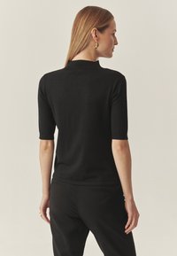 Schwarzes kurzärmliges Oberteil mit hohem Kragen, enganliegender Form und weichem Material. Kombiniert mit schwarzen, maßgeschneiderten Hosen, von hinten betrachtet.