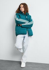 Zilā puffer jaka ar kapuci, trīs baltas svītras uz piedurknēm, ar rāvējslēdzēju priekšā un sānu kabatām; uzvalkota virs gaiši ziliem džinsi un baltām sporta kedām.