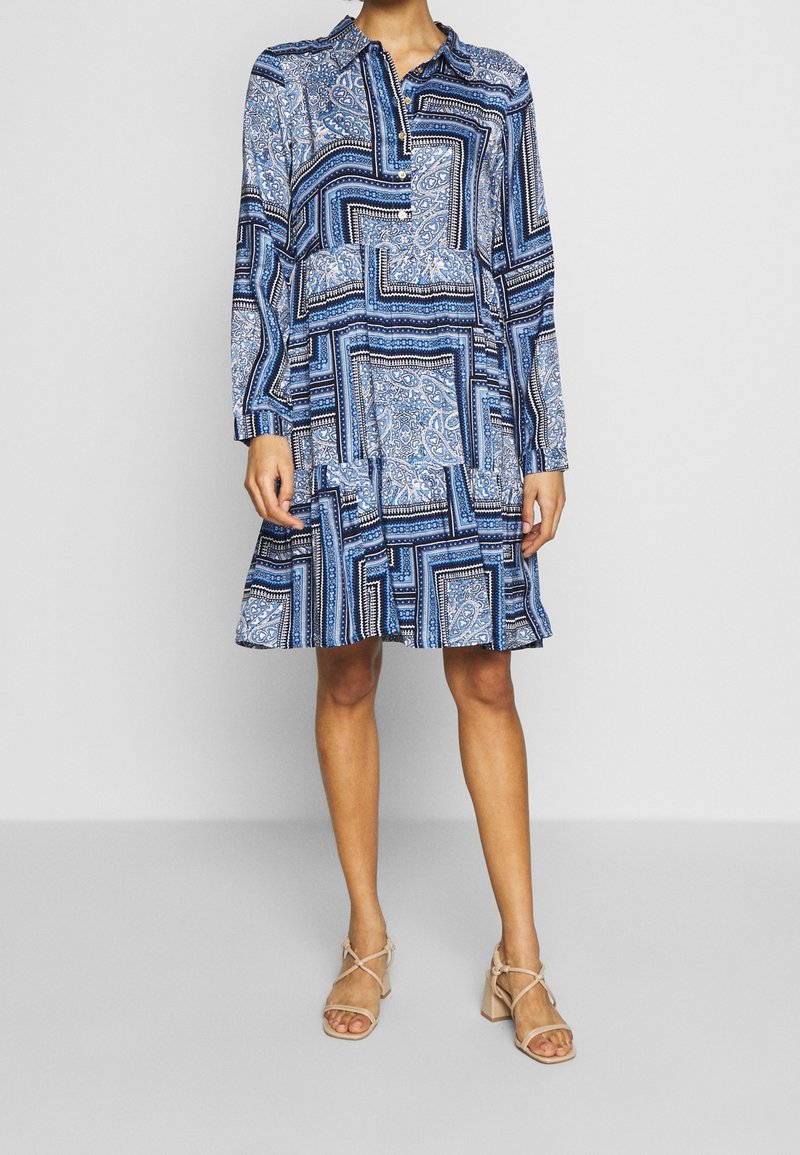 Femme portant une robe bleue à manches longues à motifs avec une jupe à volants et des sandales beiges à talons blocs et à brides, debout sur un fond uni.