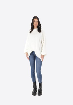 Maglione bianco di grandi dimensioni in maglia con maniche lunghe, abbinato a jeans blu aderenti e stivaletti neri chunky su uno sfondo neutro.