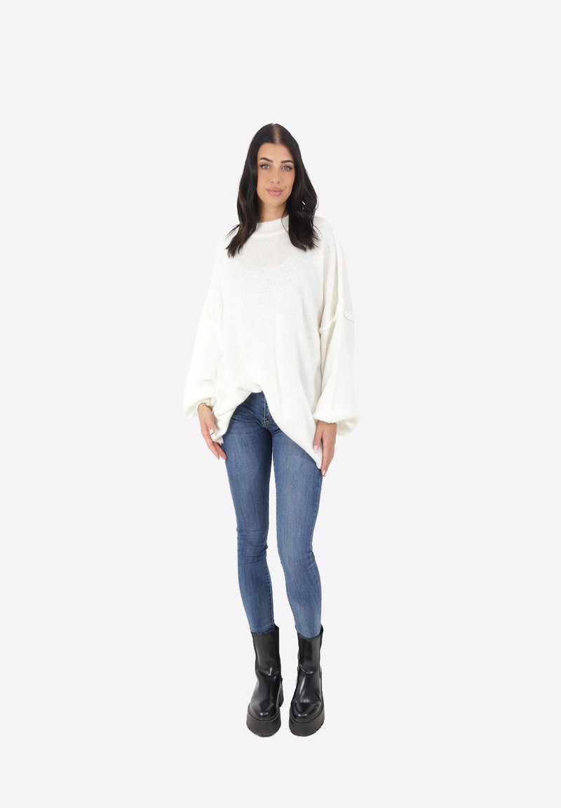 Maglione bianco di grandi dimensioni in maglia con maniche lunghe, abbinato a jeans blu aderenti e stivaletti neri chunky su uno sfondo neutro.