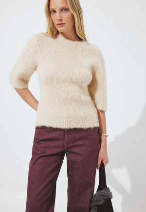 Mujer con suéter crema de peluche y bufanda gris, pantalones anchos burdeos, botas negras, sosteniendo un bolso tote negro, de pie sobre fondo blanco.