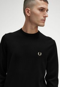 Schwarzer Strickpullover mit rundem Halsausschnitt und langen Ärmeln. Verfügt über ein kleines goldenes Logo auf der Brust. Weiche Textur ohne Muster.