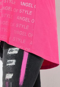 T-shirt rosa con stampa ripetuta "ANGEL OF STYLE", abbinato a jeans neri sbiaditi con macchie bianche e una striscia rosa di accento.