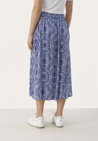 Frau in einem blau-weiß gemusterten Midikleid mit elastischem Bund und weißen Sneakers, die vor einem schlichten Hintergrund steht.