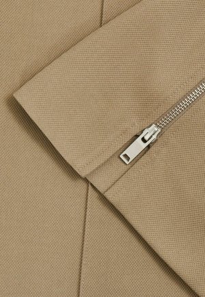 Tissu beige avec une surface texturée et une couture diagonale, présentant une fermeture éclair en argent avec un tirette rectangulaire.