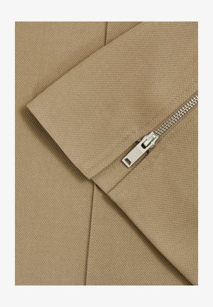 Beige kangas tekstuuriga pinnaga ja diagonaalse õmblusega, millel on hõbedane tõmblukk, millel on ristkülikukujuline tõmblemislink.