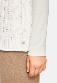 Hand resting by side wearing white cable-knit sweater and beige knitted pants, visible clothing textures and small metallic tag on sweater.