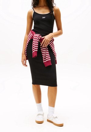 Mujer con camiseta de tirantes negra, falda midi negra, zapatillas blancas con calcetines y un suéter a rayas rojas y blancas atado a la cintura.