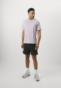 Helllila Baumwoll-T-Shirt mit Rundhalsausschnitt und kurzen Ärmeln, kombiniert mit schwarzen Shorts. Sportliche Schuhe und weiße Socken vervollständigen das Outfit.