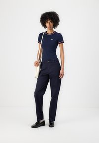 Mørkeblåtet t-skjorte med rød og hvit ringdetalj, parret med mørkeblå bukser og svarte loafers. Modell som holder en beige totebag.