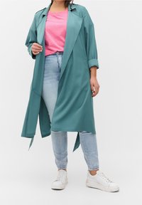 Donna che indossa un lungo cappotto turchese sopra una maglietta rosa, jeans azzurri chiari con strass vicino ai polsini e sneakers bianche, in piedi contro uno sfondo bianco.