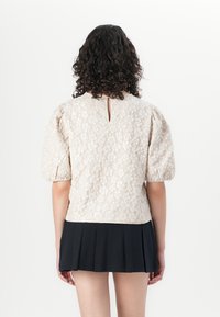 Lys beige topp med pufferm og blomstertekstur, nøkkelhull detalj bak, parret med et svart plisserte skjørt. Glatt stofffinish.