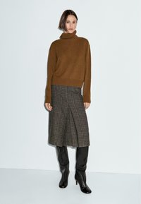 Brązowy sweter z golfem, spódnica midi w kratę z fałdami, czarne kozaki za kolano. Miękka tekstura, luźny krój i brązowo-szara paleta kolorów.