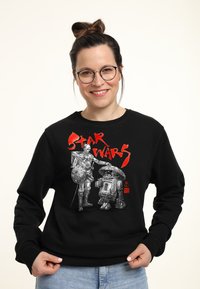 Sudadera negra con gráfico en blanco y rojo de "Star Wars" que presenta a los personajes C-3PO y R2-D2. El tejido parece suave y tiene un corte holgado.
