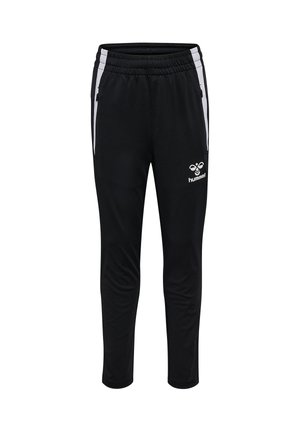 Smilende dreng står med hænderne bag ryggen iført gul og sort hummel sportjakke, sorte hummel bukser og sorte sneakers med hvide såler.