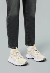 Weiße und beige Sneakers mit einer strukturierten Oberseite, Schnürsenkeln und einer grauen Außensohle. Kombiniert mit dunkler Jeans und weißen Crew-Socken.