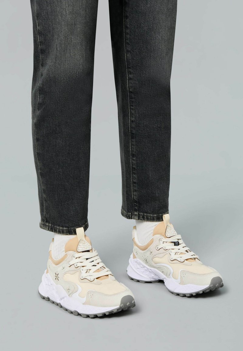 Weiße und beige Sneakers mit einer strukturierten Oberseite, Schnürsenkeln und einer grauen Außensohle. Kombiniert mit dunkler Jeans und weißen Crew-Socken.