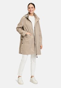 Veste longue beige, imperméable, avec une capuche, une fermeture éclair à l'avant, deux grandes poches latérales et un motif à carreaux à l'intérieur. Associée à un pantalon blanc et des baskets.