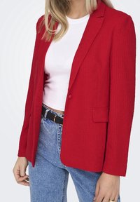 Rød tekstureret blazer med notch-revers, enkelt knaplukning og to forlommer, kombineret med en hvid top og højtsiddende denimjeans.