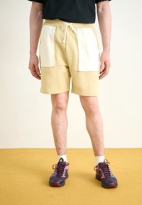 Lys beige shorts med hvite lommeposter, snøring i livet, kombinert med flerfargede joggesko. Mot en gul gulvbakgrunn.