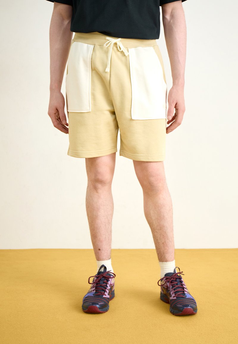 Lys beige shorts med hvite lommeposter, snøring i livet, kombinert med flerfargede joggesko. Mot en gul gulvbakgrunn.