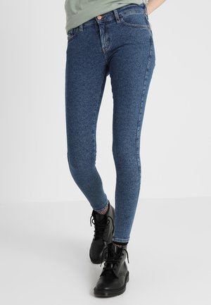Frau trägt blaue Slim-Fit-Jeans, Netzstrümpfe und schwarze Schnürknöchelstiefel, steht mit einem Bein vor dem anderen verschränkt.
