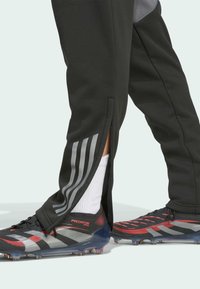 Schwarze Sporthosen mit seitlichen Reißverschlüssen und grauen Streifen, kombiniert mit schwarzen und roten Fußballschuhen mit texturiertem Obermaterial und transparenten Sohlen.