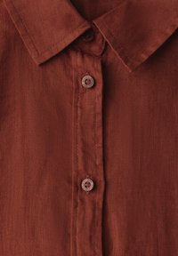 Rust-colored linen shirt featuring a classic collar, five buttons, and a subtle textured surface. Buttons are round with a stamped design.