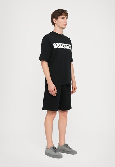T-shirt noir avec "OBSESSED" en lettres blanches en gras, associé à un short noir. Le mannequin porte des baskets en daim gris; design minimaliste.