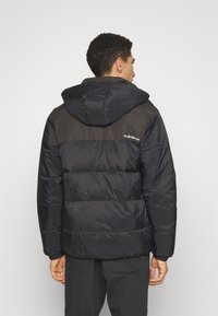 Black puffer jacket with a hood, featuring contrasting dark gray shoulder panels. Thick quilted texture and logo detail on the back.