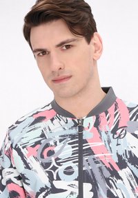Jachetă bomber multicolor cu modele abstracte în culorile roz, albastru și gri; închidere cu fermoar în față și guler gri, cu textură din țesătură.