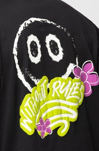 Tissu noir avec un visage de smiley blanc aux bords irréguliers, texte jaune vif "WITHOUT RULES" et deux fleurs de dessin animé violettes.