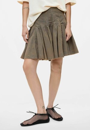 Mujer con falda plisada verde oliva, camiseta de manga corta color crema y zapatos planos negros de malla con cordones, de pie sobre un fondo blanco.