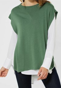 Zelený oversized svetr s kulatým výstřihem, krátkými rukávy, nahoře přes dlouhý bílý košili. Jednoduchá textura, ležérní design.