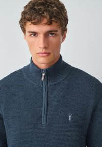 Navy-blau gestrickter Pullover mit hohem Kragen und halbem Reißverschluss. Strukturiertes Gewebe und kleines Logo-Detail auf der Brust.