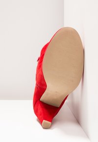 Botte rouge en suède à talon haut reposant sur le côté, montrant une semelle beige lisse et un talon court effilé sur un fond blanc d'angle.