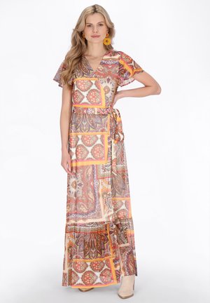 Kvinde iført en farverig, mønstret kortærmet omslagsmaksikjole med orange øreringe og ankelstøvler, stående med den ene hånd på hoften.