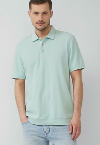 Jasnozielona koszulka polo o teksturowanej tkaninie, z trzema guzikami i krótkimi rękawami, w zestawie z niebieskimi dżinsami.