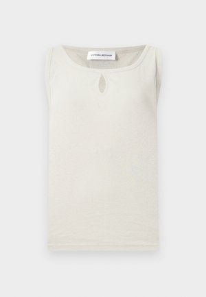 Vrouw met donker haar, gekleed in een beige mouwloos topje, witte wijde broekspijpen, zwarte riem en zwarte open teen hakken, staand tegen een witte achtergrond.