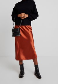 Zwarte gebreide trui, oranje satijnen midirok, zwarte enkellaarzen en een kleine zwarte handtas met kralen en een bandje. Textuur- en gladde elementen.